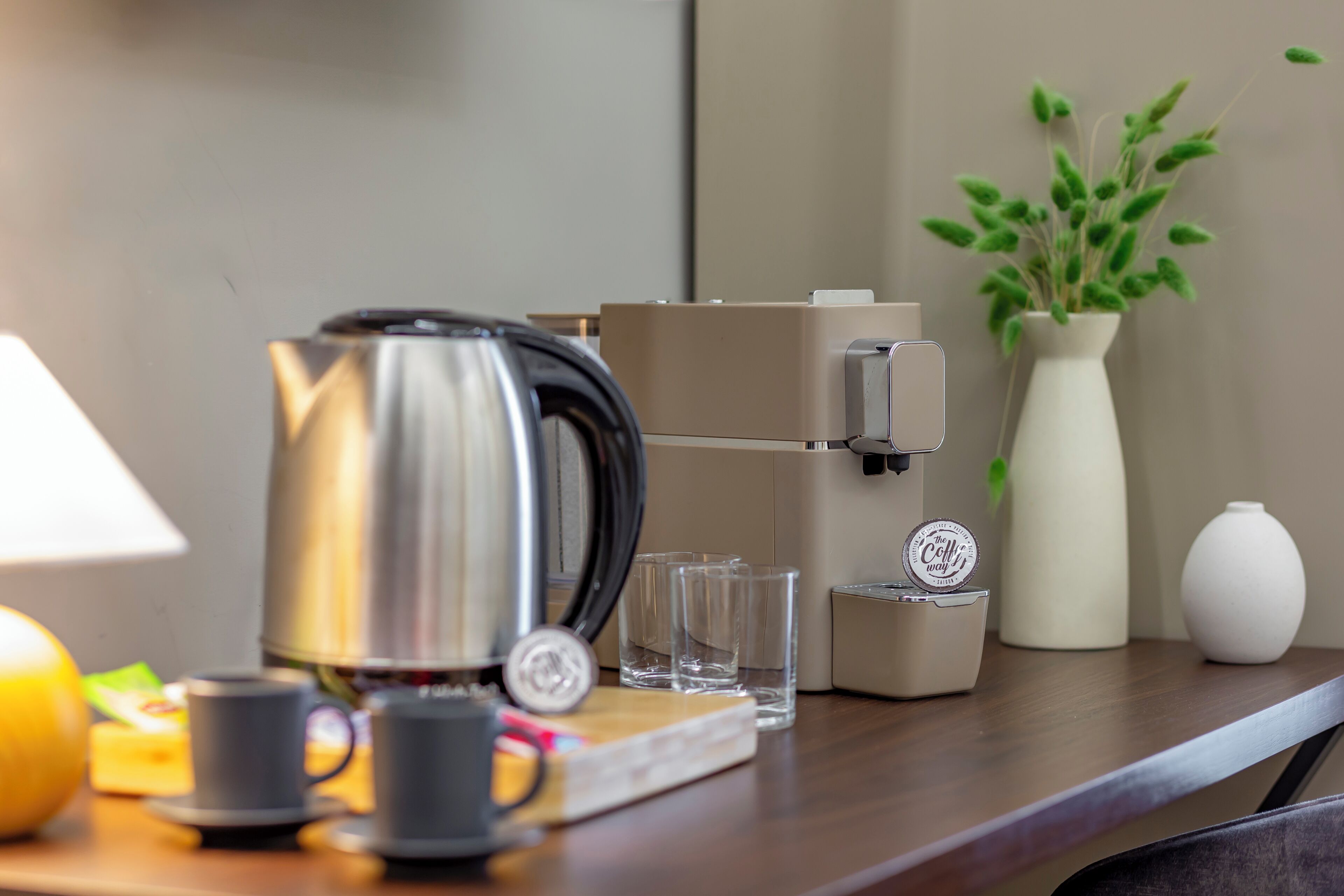 Uma área de cozinha com cafeteira, chaleira, um vaso com plantas verdes e uma máquina de café em uma bancada de madeira.