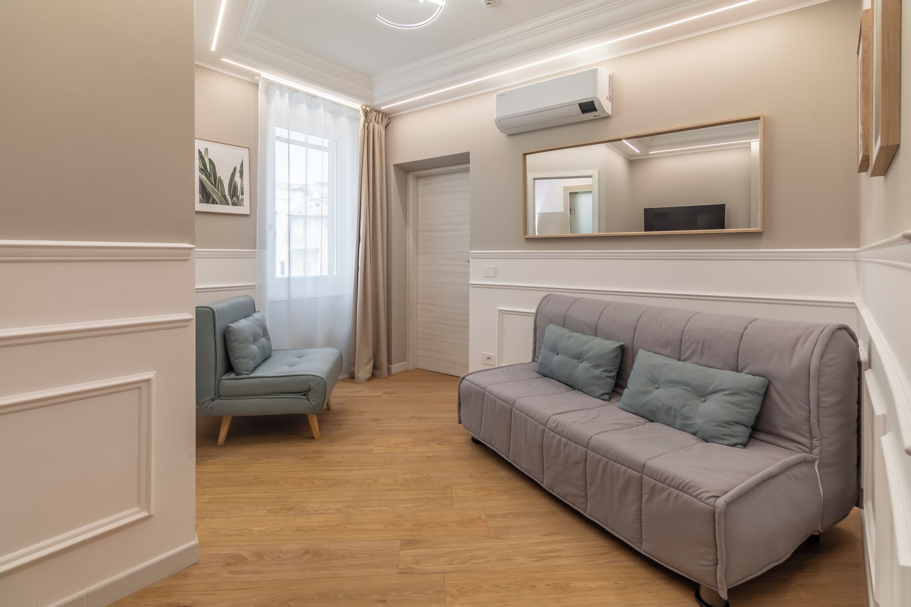 A room with a beige sofa, a grey armchair, a wooden floor, a large mirror, and an air conditioning unit.