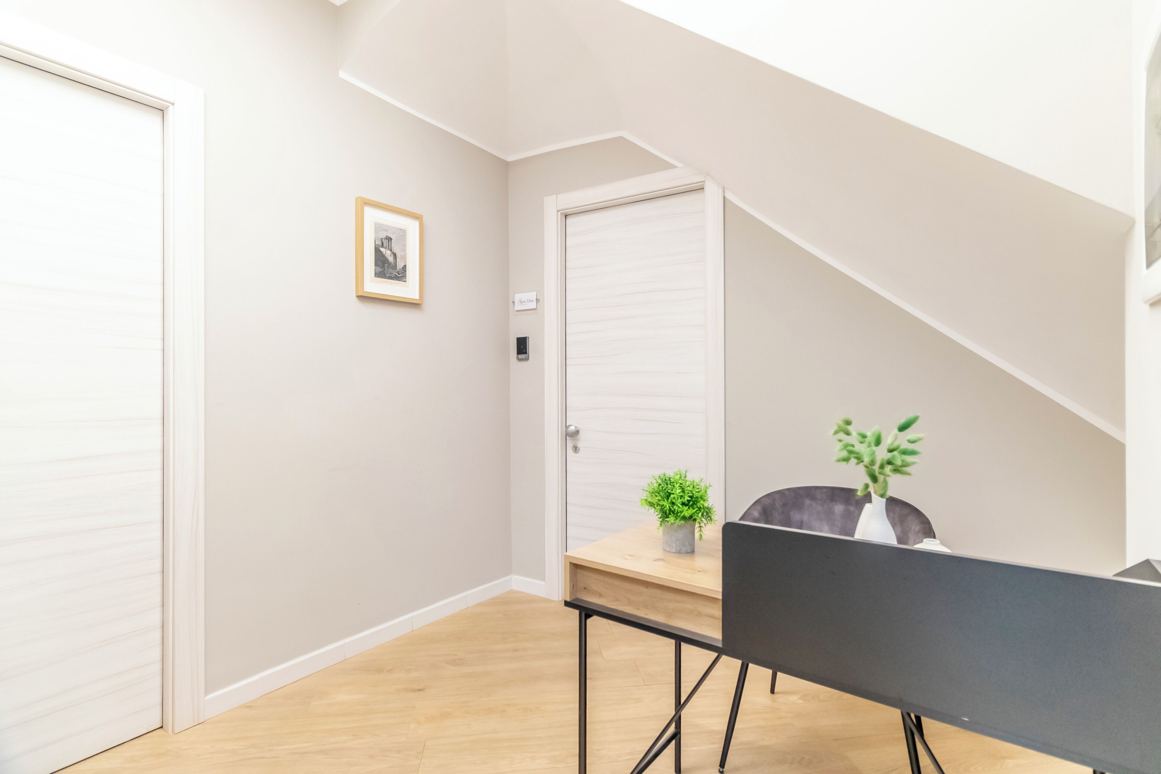 A room with a wooden floor, a black chair, a small table with a plant, and a framed picture on the wall.