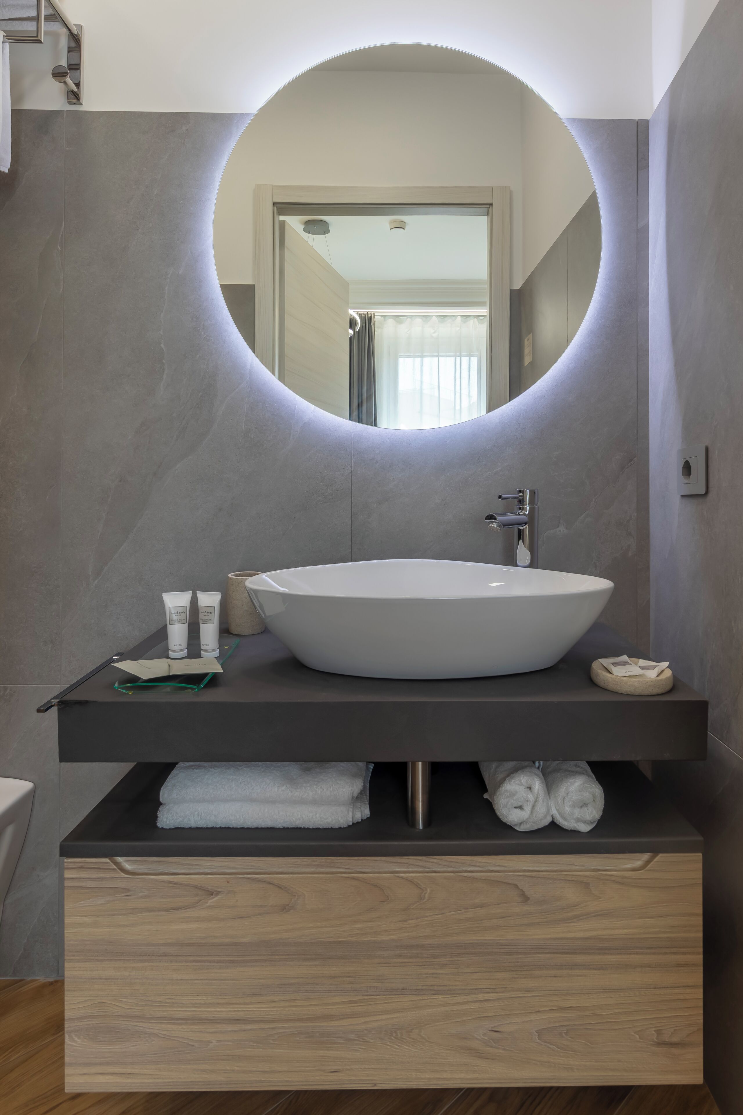 Un baño moderno con un espejo redondo, un lavamanos blanco tipo recipiente y un mueble de tocador de madera con un perchero.