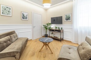 A living room with a beige sofa, a round table, a TV, and framed artwork on the walls.