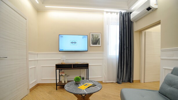 Een woonkamer met een flatscreen televisie, een ronde marmeren tafel, een grijze bank en een kleine plant.