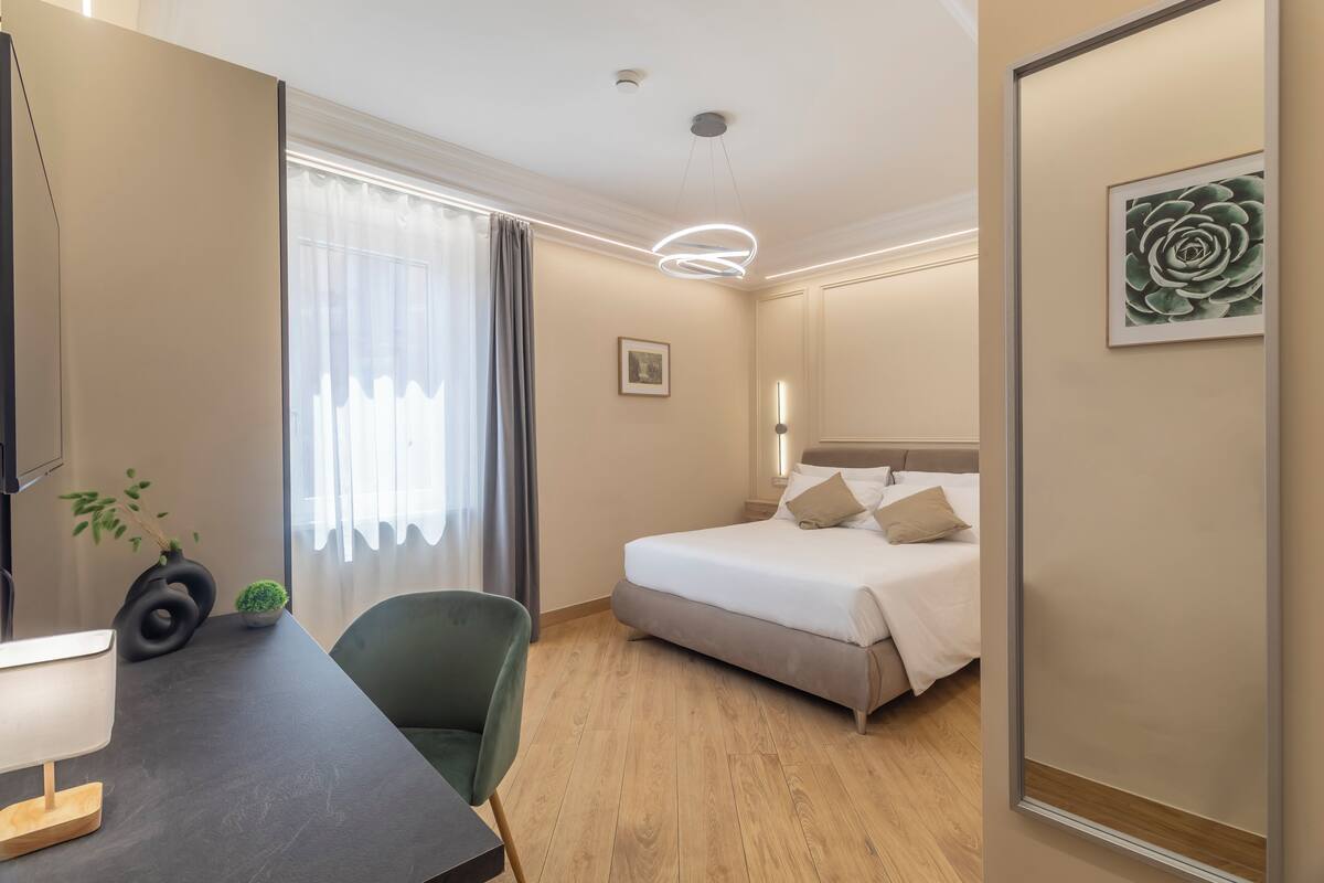 Una habitación de hotel con una cama, un escritorio con lámpara, una silla, una planta y un cuadro en la pared.
