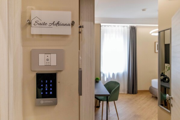 A hotel room with a digital keypad lock, a sign indicating 'Suite Adriana,' a dining area with a table and chairs, and a view of a window with curtains. - Mom Apartment Rooms&Suites (Tivoli)