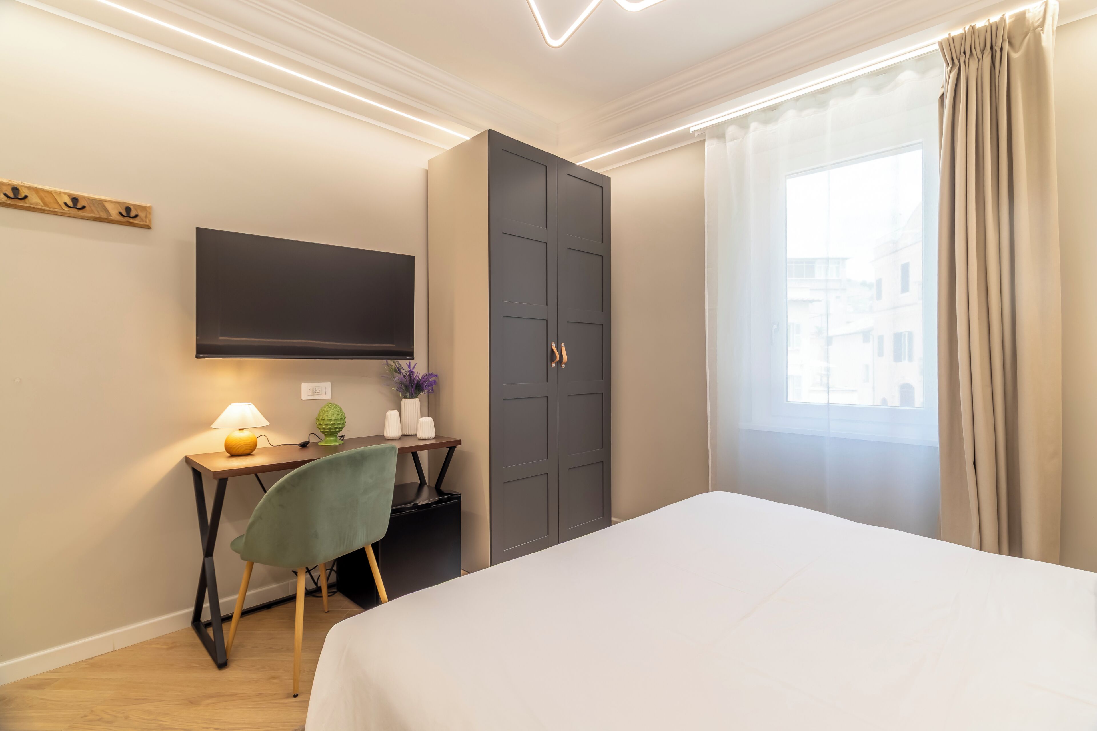 A hotel room with a bed, a desk with a lamp, a chair, a wardrobe, and a TV mounted on the wall.