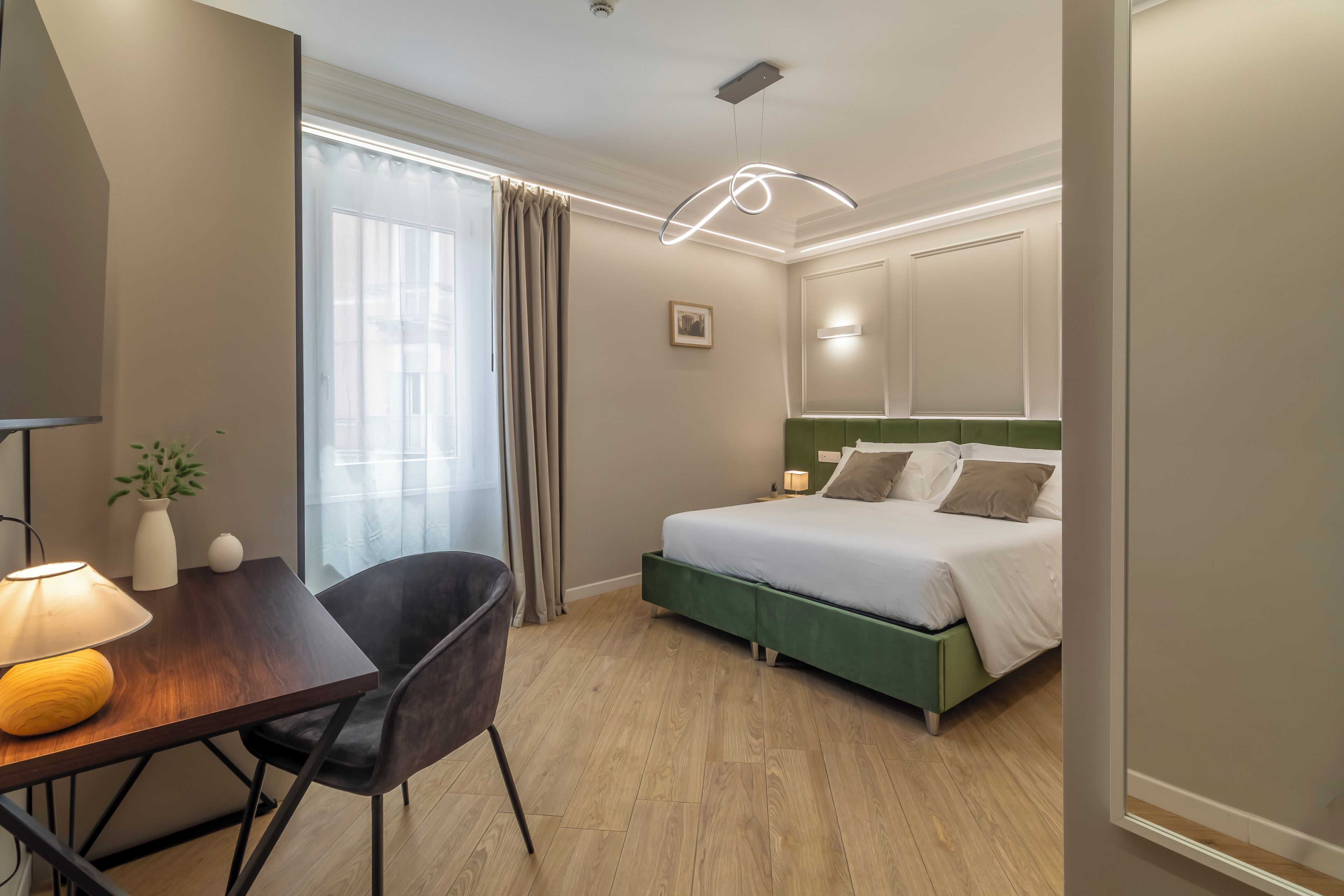 Ein Hotelzimmer mit einem Bett, einem Schreibtisch mit Lampe, einem Sessel, einem Fenster mit Vorhängen und einer modernen Leuchte.