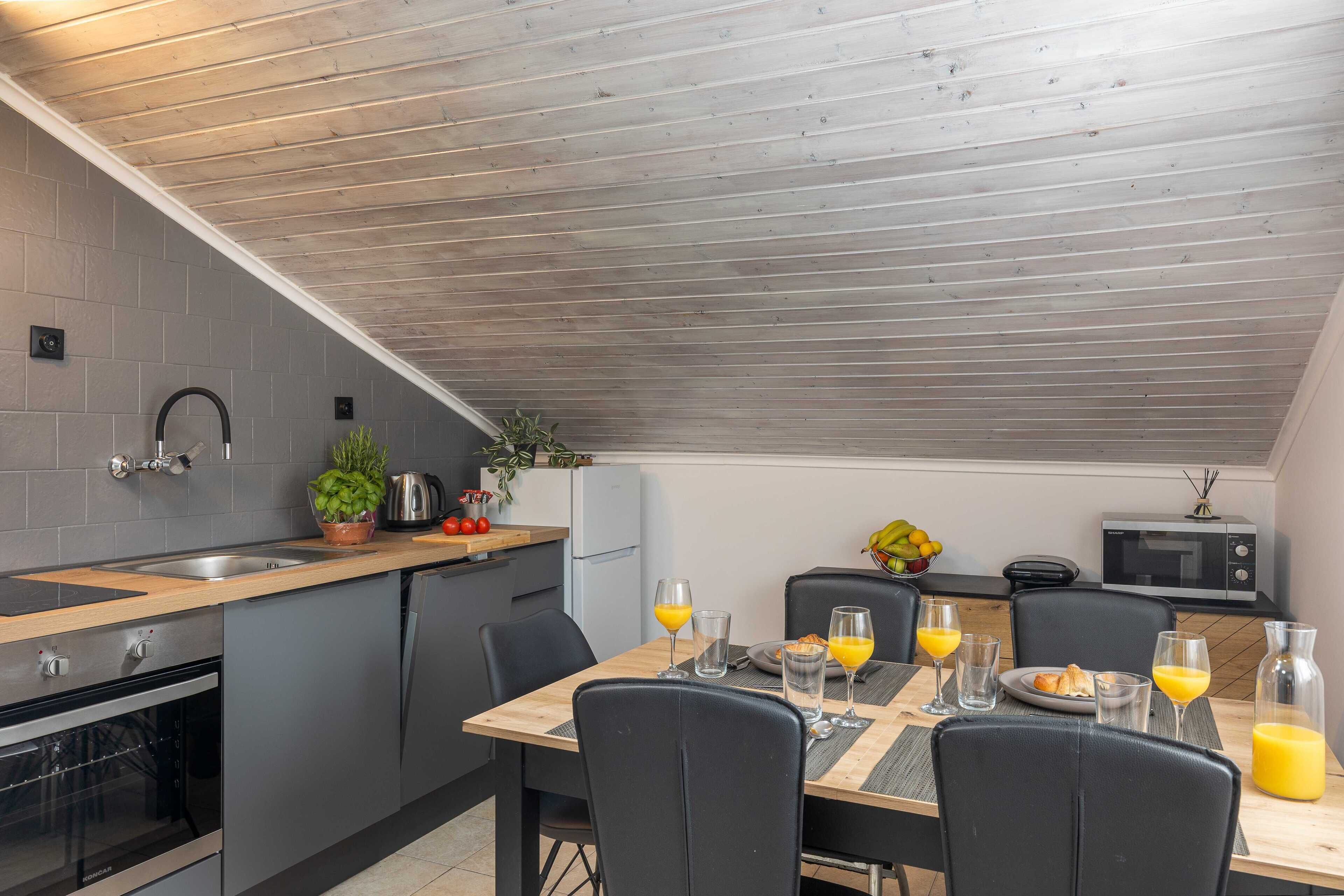 A modern kitchen with grey tiles, wooden ceiling, stainless steel appliances, and a dining area with black chairs and breakfast setup.