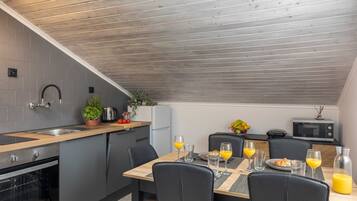 A modern kitchen with grey tiles, wooden ceiling, stainless steel appliances, and a dining area with black chairs and breakfast setup.