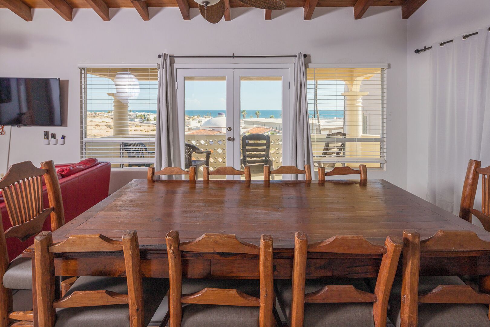 Un espace repas comprenant une grande table et des chaises en bois, un canapé rouge et une vue sur la mer grâce à de grandes fenêtres.