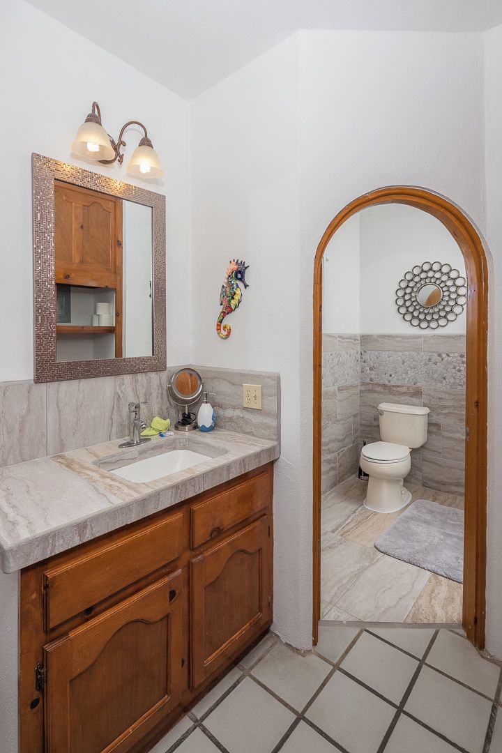 A bathroom with a marble sink, wooden cabinets, a mirror with wall sconces, a decorative seahorse, and a toilet with a marble backsplash.
