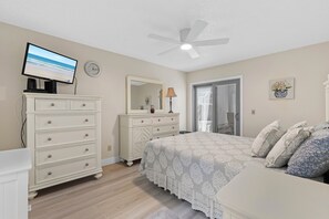 Une chambre à coucher comprenant un lit, une commode, un téléviseur, un ventilateur de plafond et une porte donnant sur un balcon.