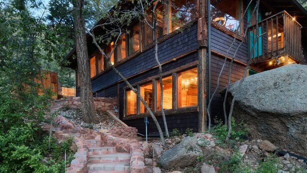 A multi-story wooden cabin with a staircase leading to the entrance, surrounded by natural vegetation and rocks. - Cabin with Pikes Views, Fireplace, Walk to Wine (El Paso County)