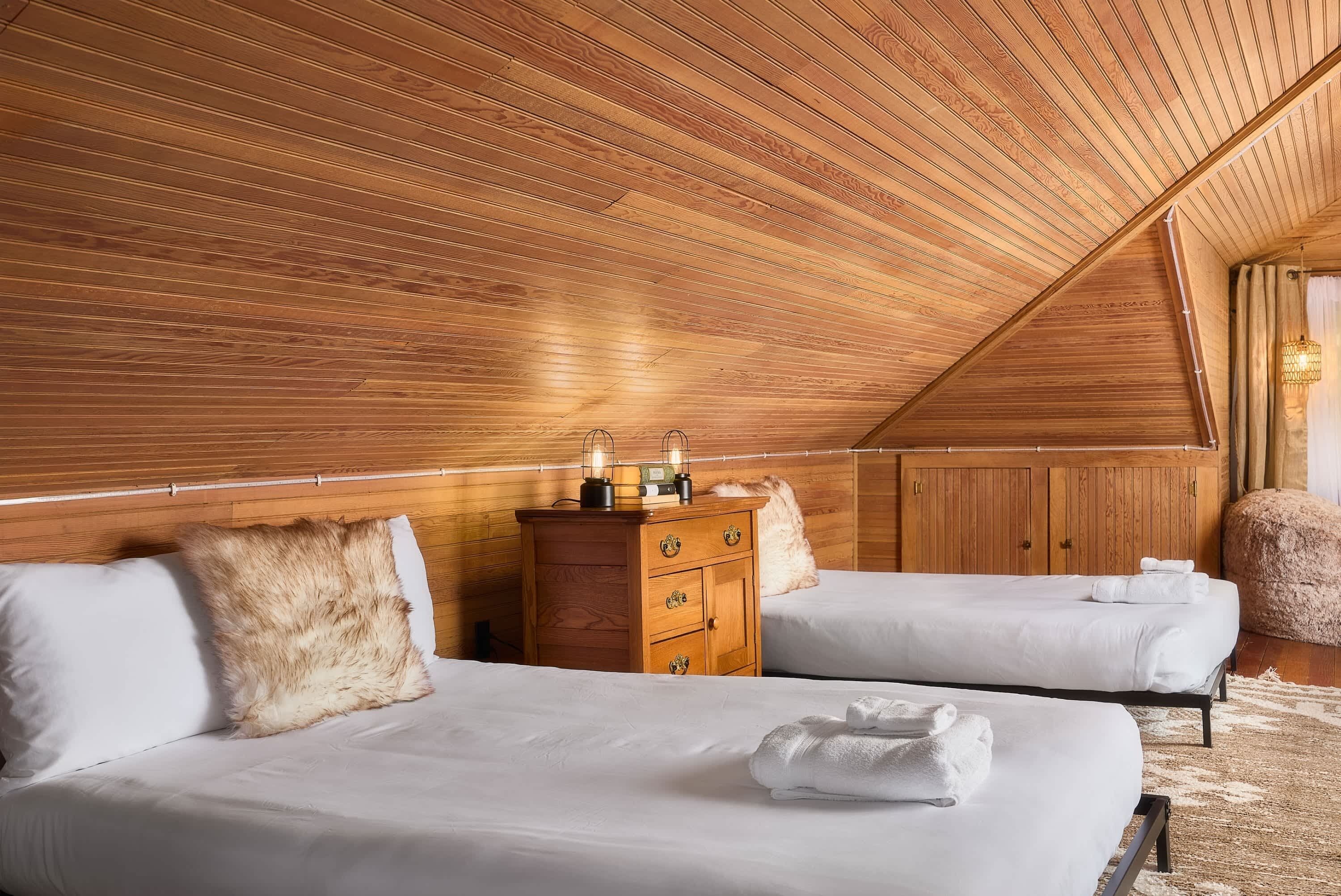 A room with wooden slatted ceiling, two beds with white bedding, a wooden dresser, and a fluffy pillow.
