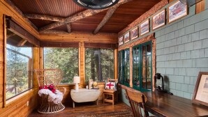 A rustic cabin interior with wooden walls, a wooden ceiling, a rocking chair, a small table, and a window with a view of trees. - Cabin with Pikes Views, Fireplace, Walk to Wine (El Paso County)