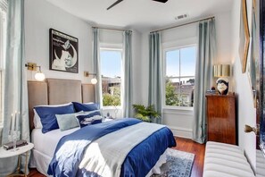 A bedroom with a large bed, two windows with curtains, a nightstand with a lamp, a framed picture on the wall, and a view of the outside. - Contemporary 4BR Victorian, Near Forsyth Park (Savannah)