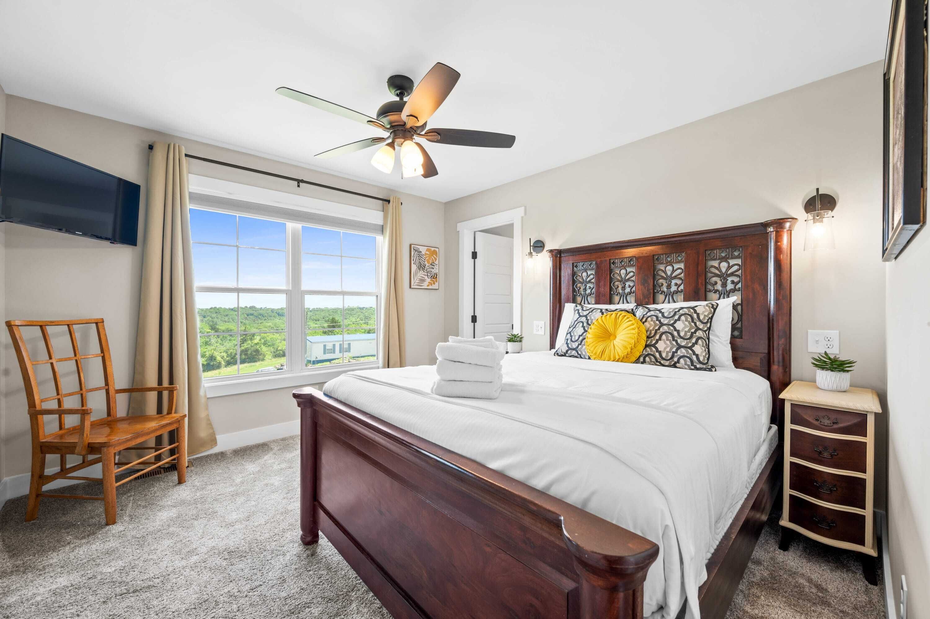 A bedroom with a large bed, a ceiling fan, a TV mounted on the wall, a window with curtains, and a view of greenery outside.