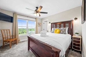 Un dormitorio con una cama grande, ventilador de techo, televisor montado en la pared, ventana con cortinas y vistas a zonas verdes.