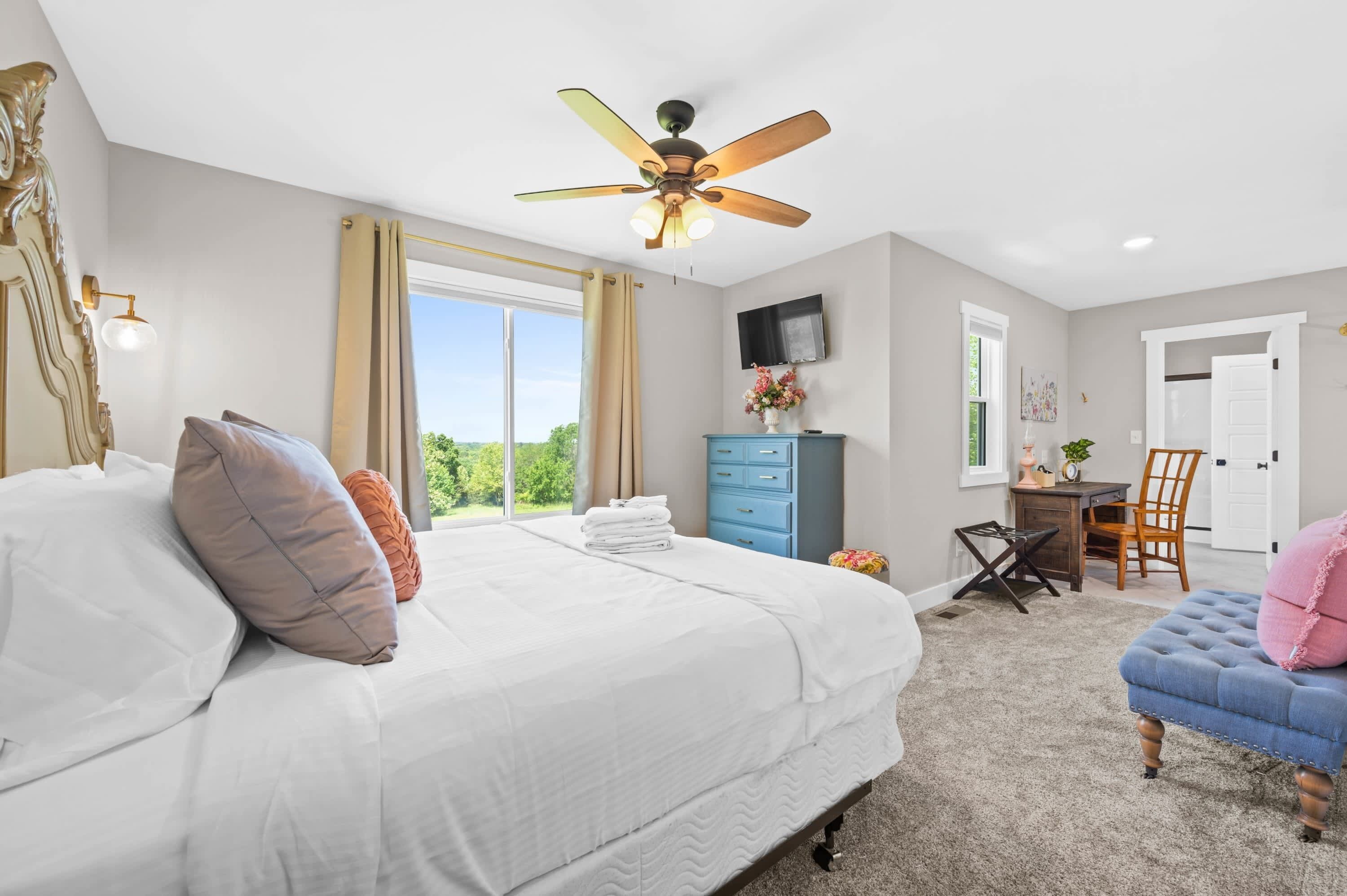 A bedroom with a large bed, a ceiling fan, a desk, a chair, and a view of the outdoors.