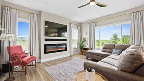 Ein geräumiges Wohnzimmer mit Kamin, Deckenventilator und großen Fenstern mit Blick ins Freie.