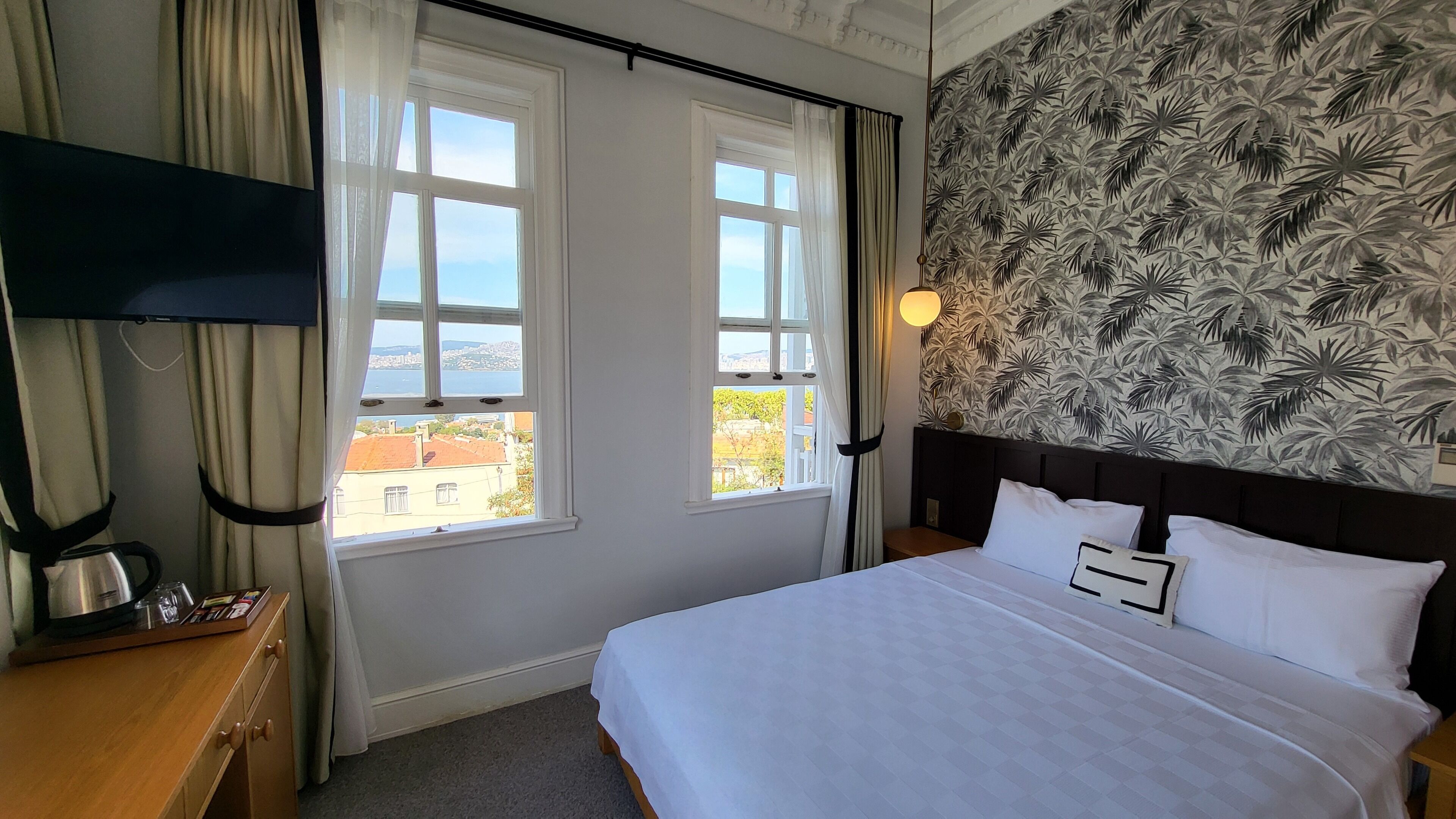 Una habitación de hotel con dos ventanas, una cama con ropa blanca, una mesita de noche, una lámpara y una pared con un diseño de hojas tropicales.