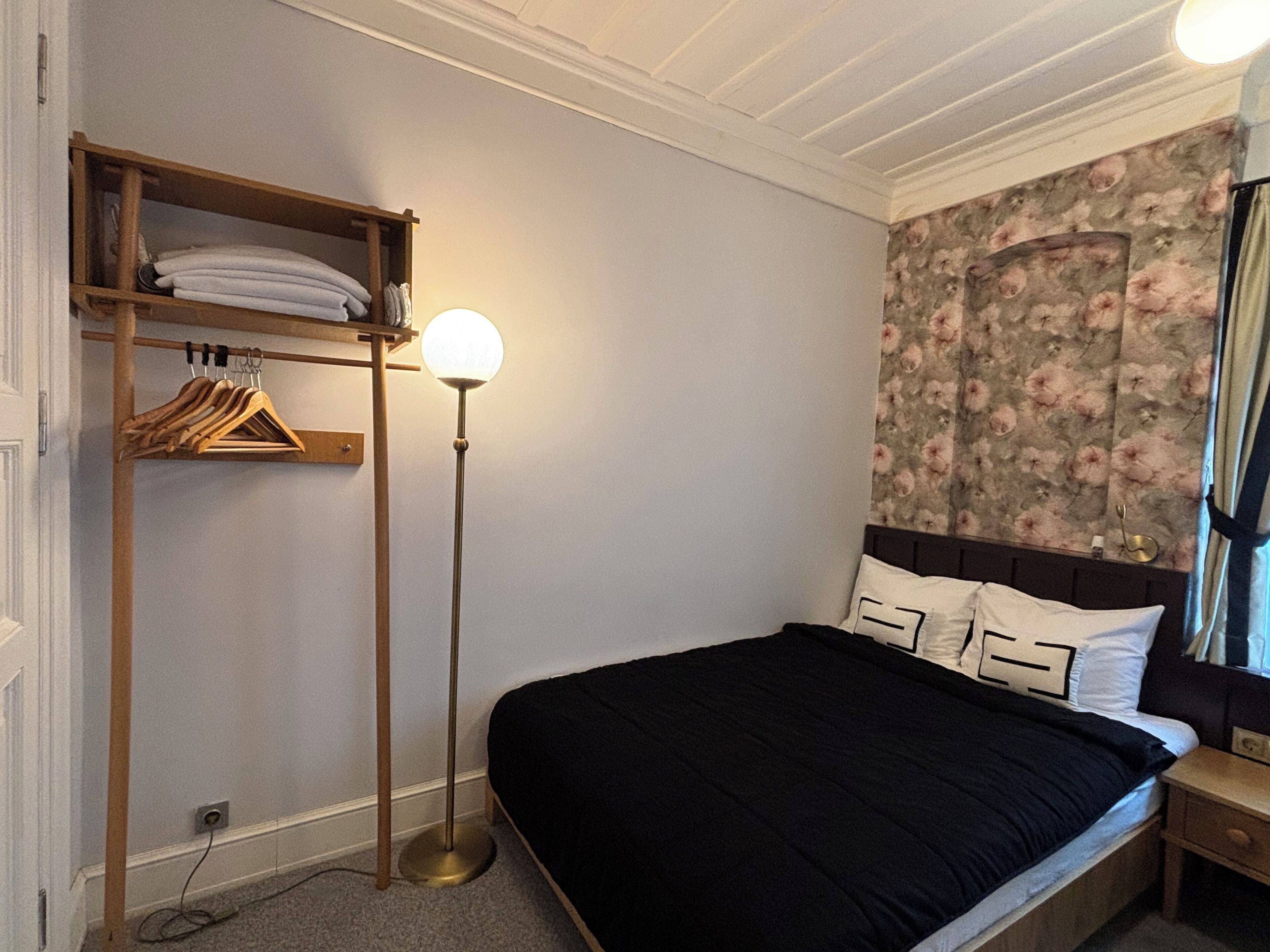 Un dormitorio con una cama, un estante de madera con perchas, una lámpara de pie y un papel tapiz floral.