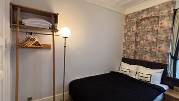 Un dormitorio con una cama, un estante de madera con perchas, una lámpara de pie y un papel tapiz floral.