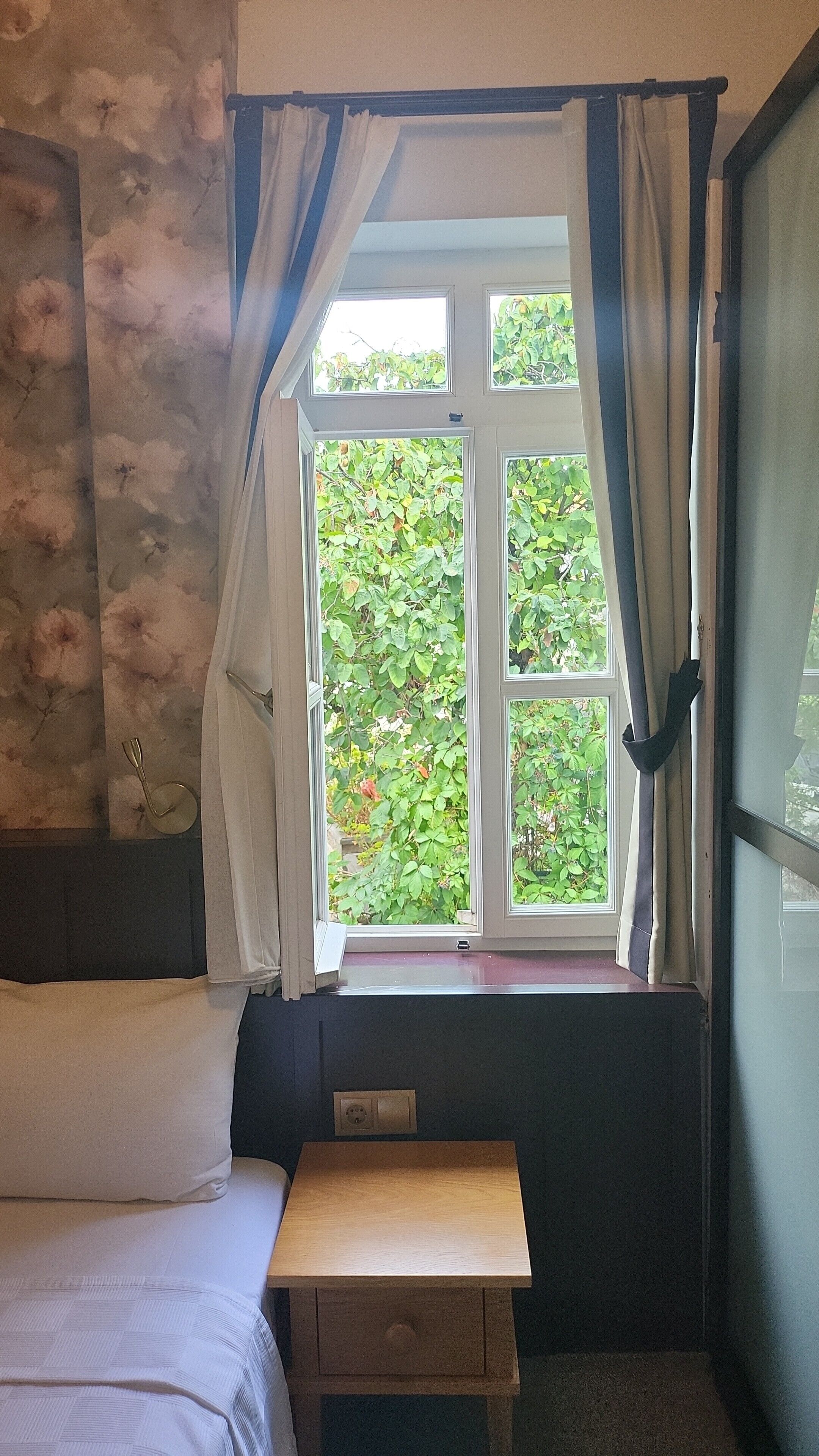 A bedroom with a bed, a small wooden nightstand, a window with curtains, and a wall with a floral pattern.
