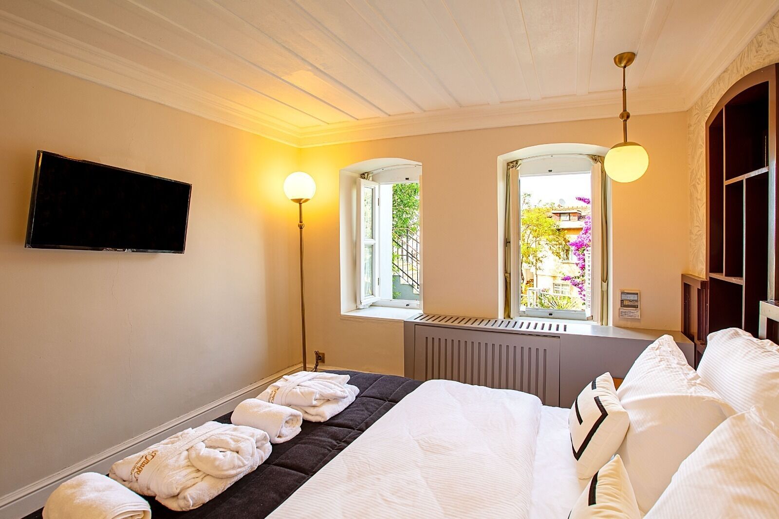 Ein Hotelzimmer mit einem Bett, zwei Fenstern, einer Heizung, einem Flachbildfernseher und einer Stehlampe.
