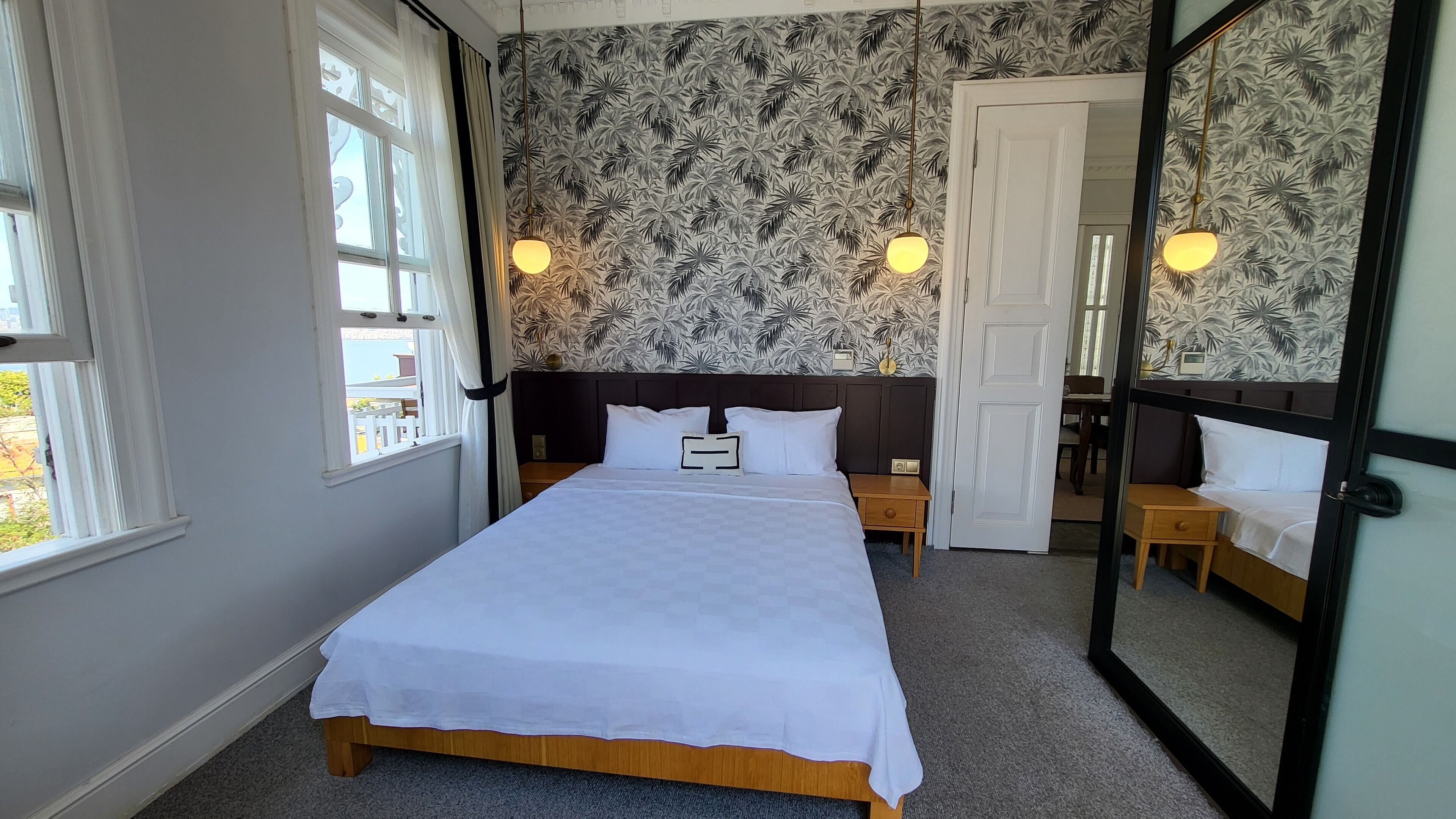 Ein ordentlich bezogenes Bett in einem Zimmer mit gemustertem Wandpapier, einem großen Spiegel und Blick nach draußen durch ein Fenster.