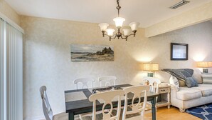 A dining area with a wooden table and white chairs, a chandelier, a painting on the wall, and a sofa with a throw blanket. - Convenient St Simons Island Home - 3 Mi to Beach! (St. Simons Island)