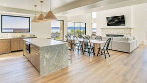 A modern kitchen with a large island, dining area with chairs, a fireplace, and large windows with a view. - Luxe Retreat w/ Theater Room: Walk to Bear Lake! (Garden City)