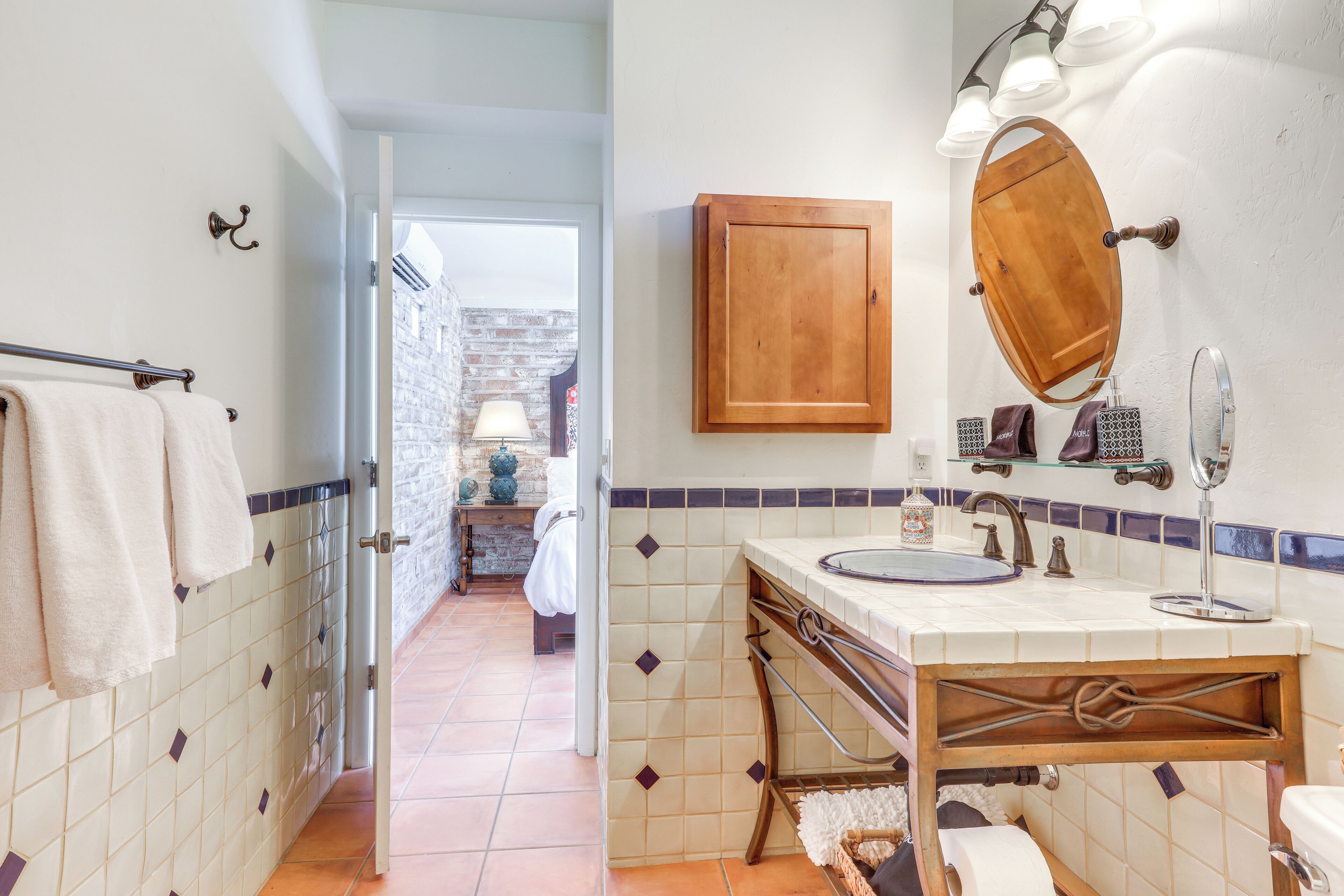 A bathroom with a wooden vanity, a mirror, and a sink. There is a towel rack with towels, a shower area visible through an open door, and a tiled floor.