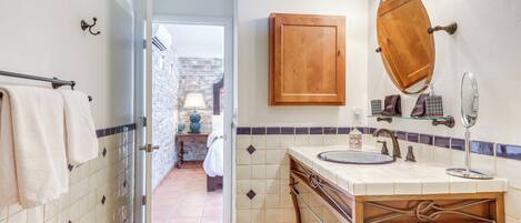 A bathroom with a wooden vanity, a mirror, and a sink. There is a towel rack with towels, a shower area visible through an open door, and a tiled floor.