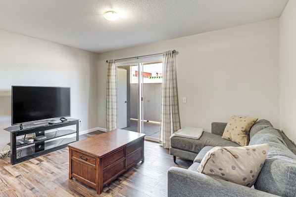A living room with a flat-screen TV on a wooden stand, a grey sofa with patterned cushions, and a wooden coffee table.