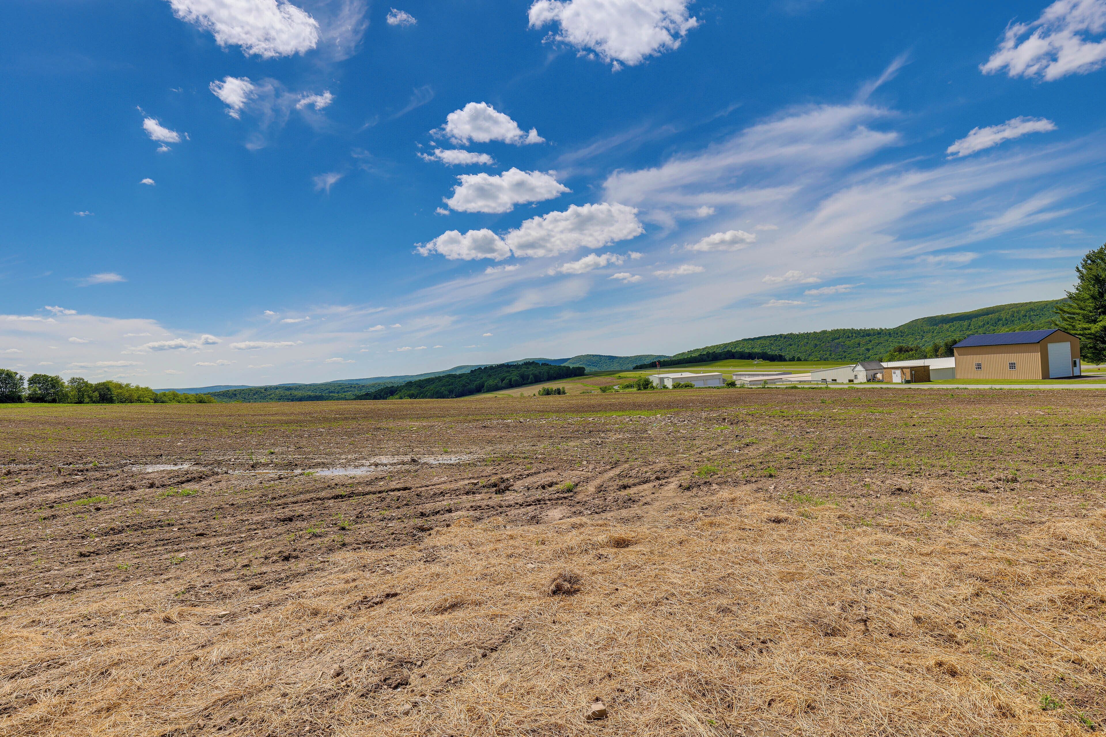 A rural landscape with a large open field, a building with a blue roof, and rolling hills in the background.