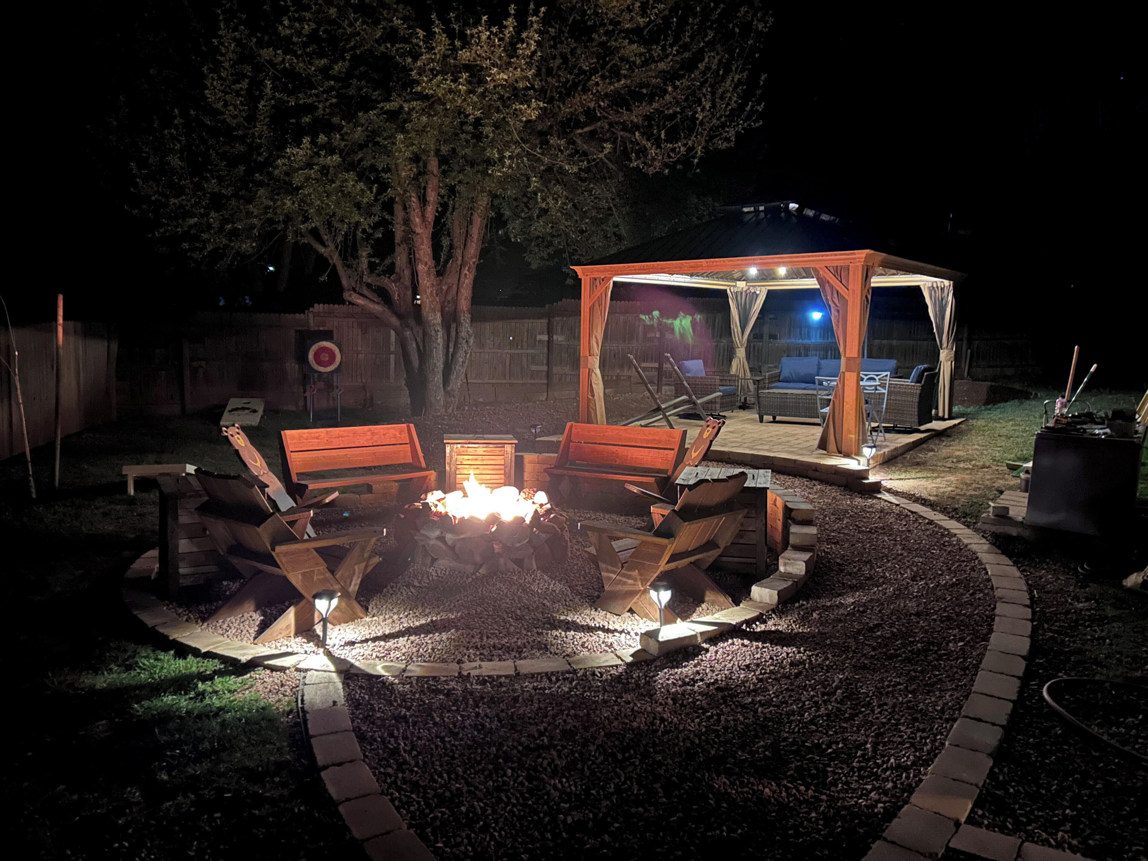 A fire pit with seating, a covered area, and a tree in a backyard setting.