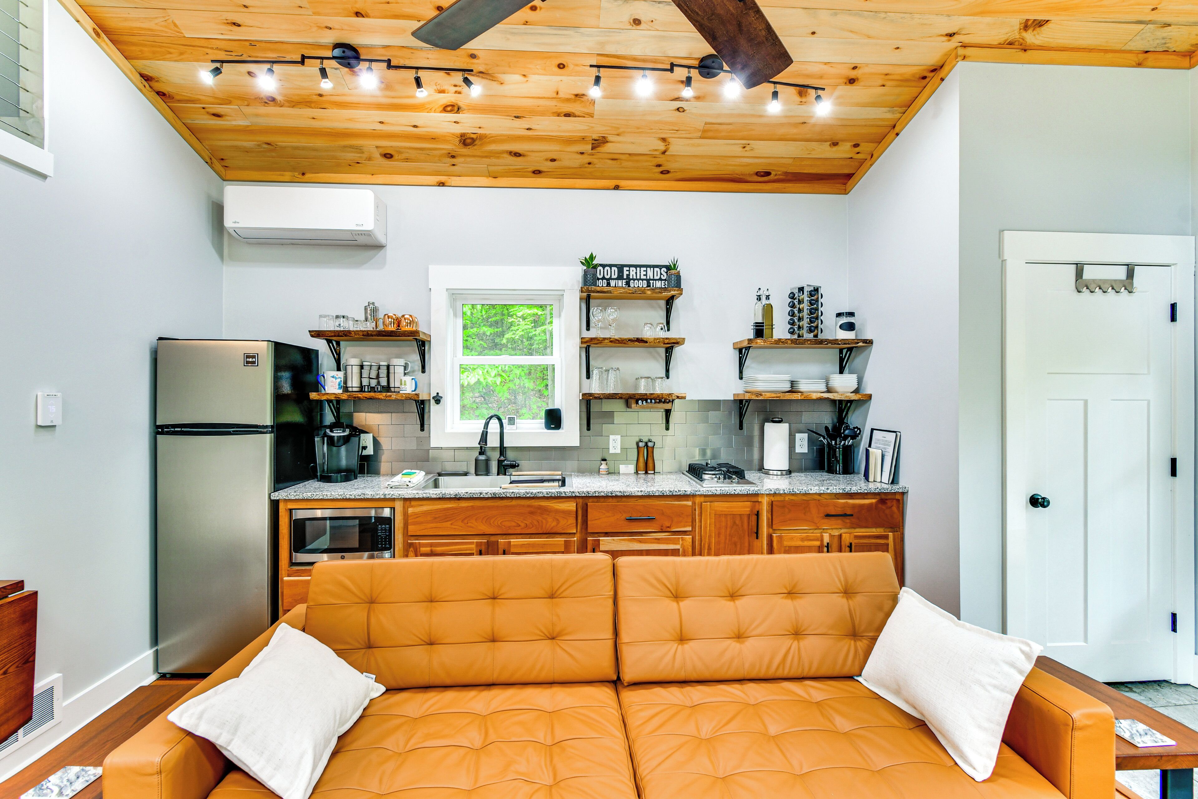 Cocina compacta con refrigerador, microondas y gabinetes de madera. Se observa una ventana con vistas a vegetación, un lavamanos y una encimera con varios objetos.
