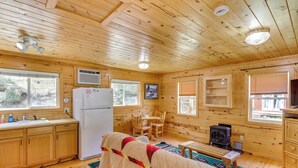 A cabin with wooden walls, a kitchen with a refrigerator, a dining table with chairs, a fireplace, and a large window. - Pets Welcome: Cozy Cabin w/ Deck in Deadwood! (Deadwood)