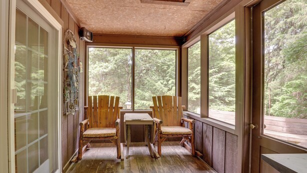 Un porche de madera con dos sillas y una mesa, ventanas amplias y vista a árboles.