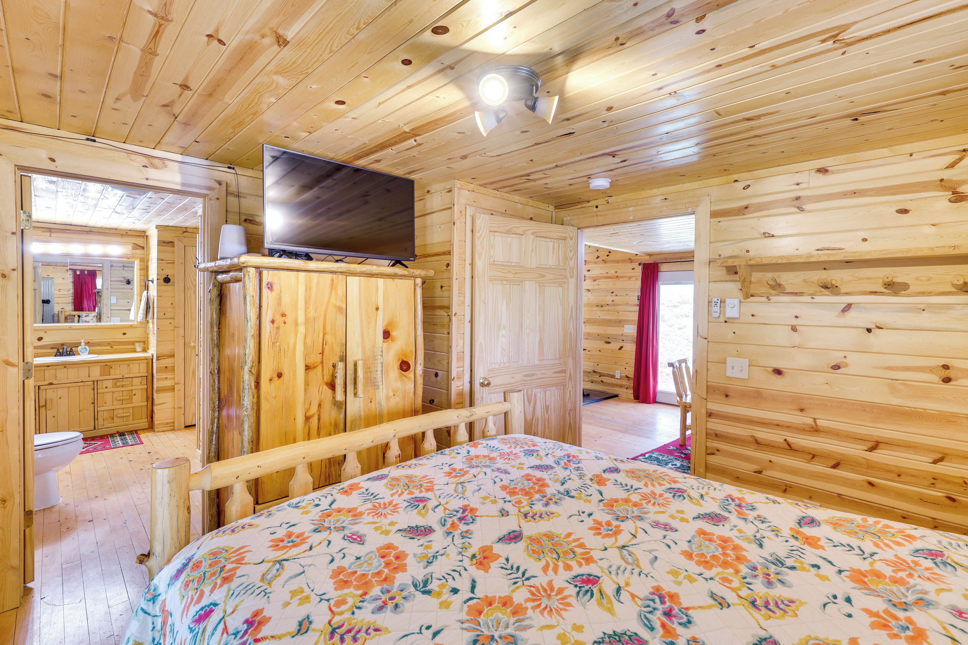 A wooden cabin room with a bed, a television on a cabinet, a bathroom with a mirror, and a door leading to another room.