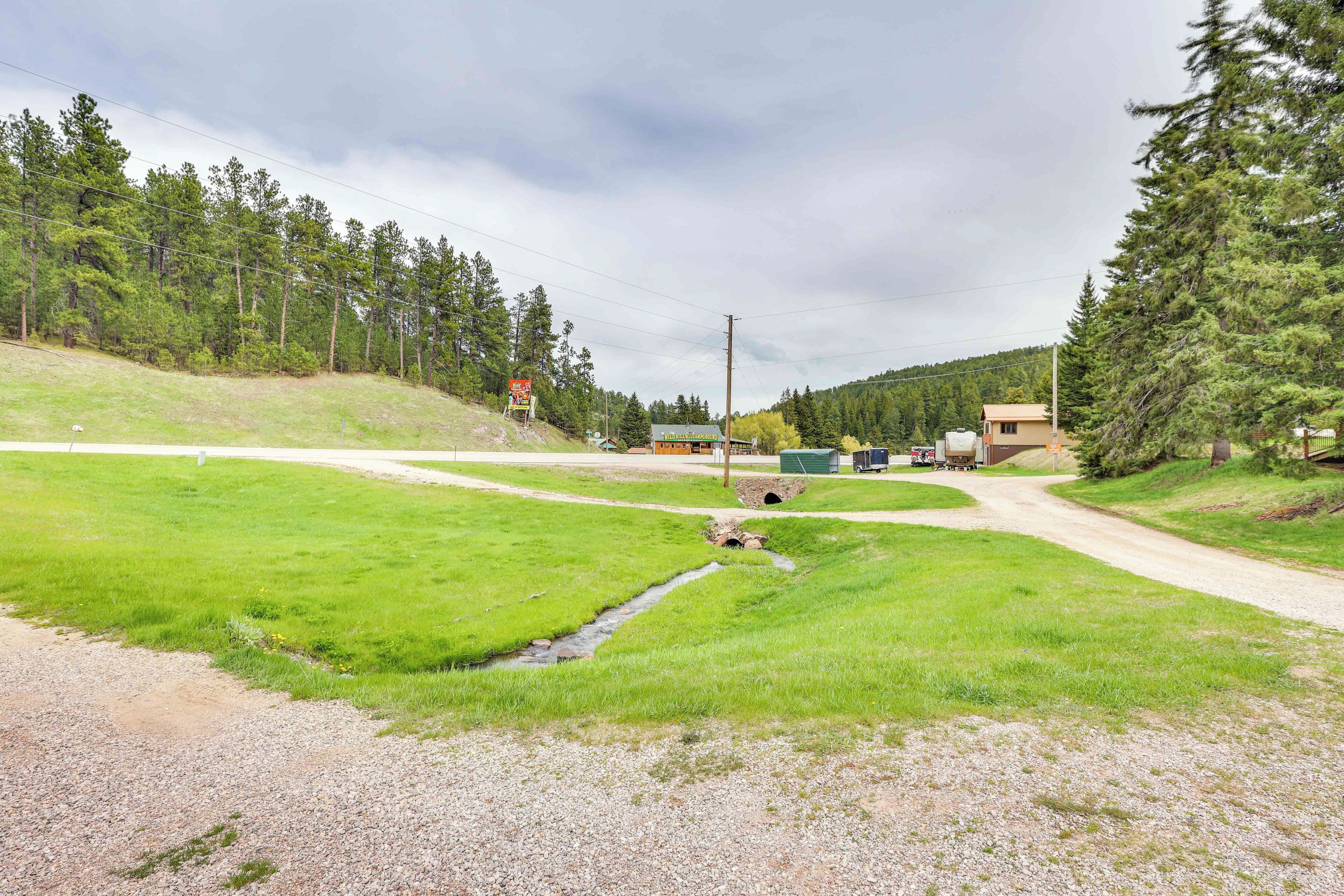 A rural road intersection with a small stream, surrounded by trees and a building with a sign.