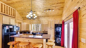 A wooden cabin interior with a kitchen, bar stools, a washing machine, and a large window. - Hike, Unwind & Bring Your Pet: Deadwood Cabin! (Deadwood)