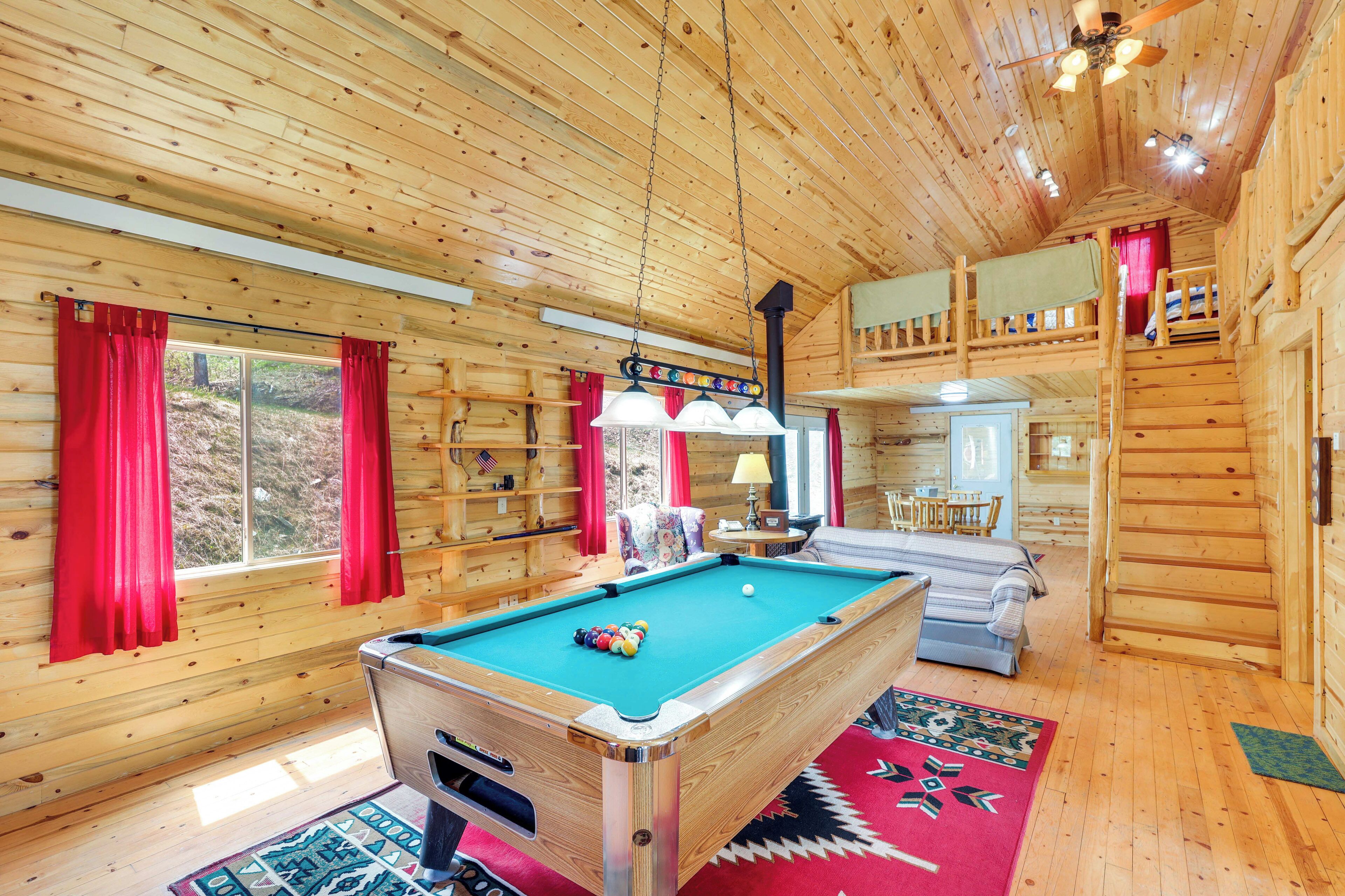 A wooden cabin interior with a pool table, red curtains, and a cozy seating area.