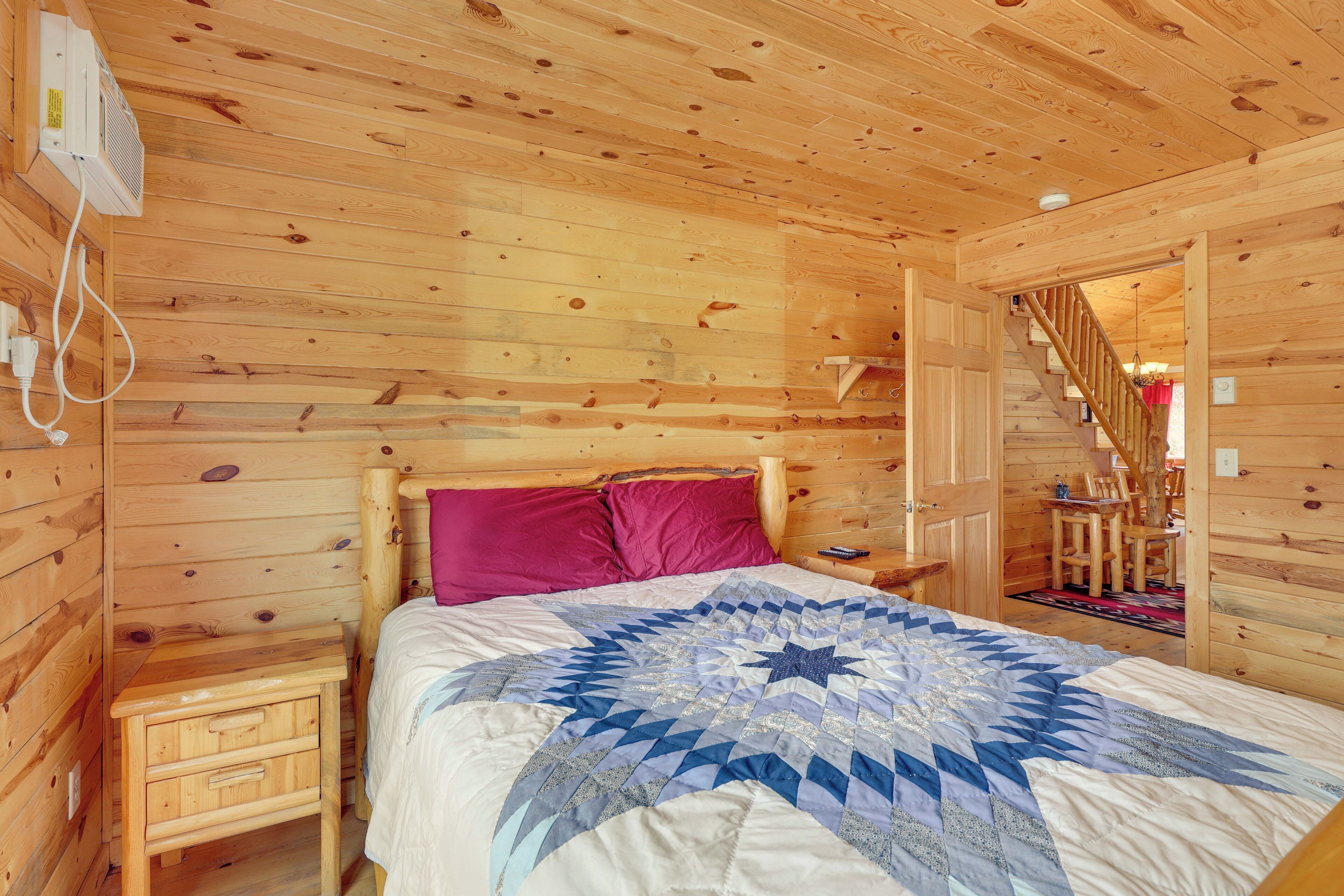 A wooden cabin bedroom with a bed, a nightstand, a red pillow, a quilt, and a staircase leading to another room.