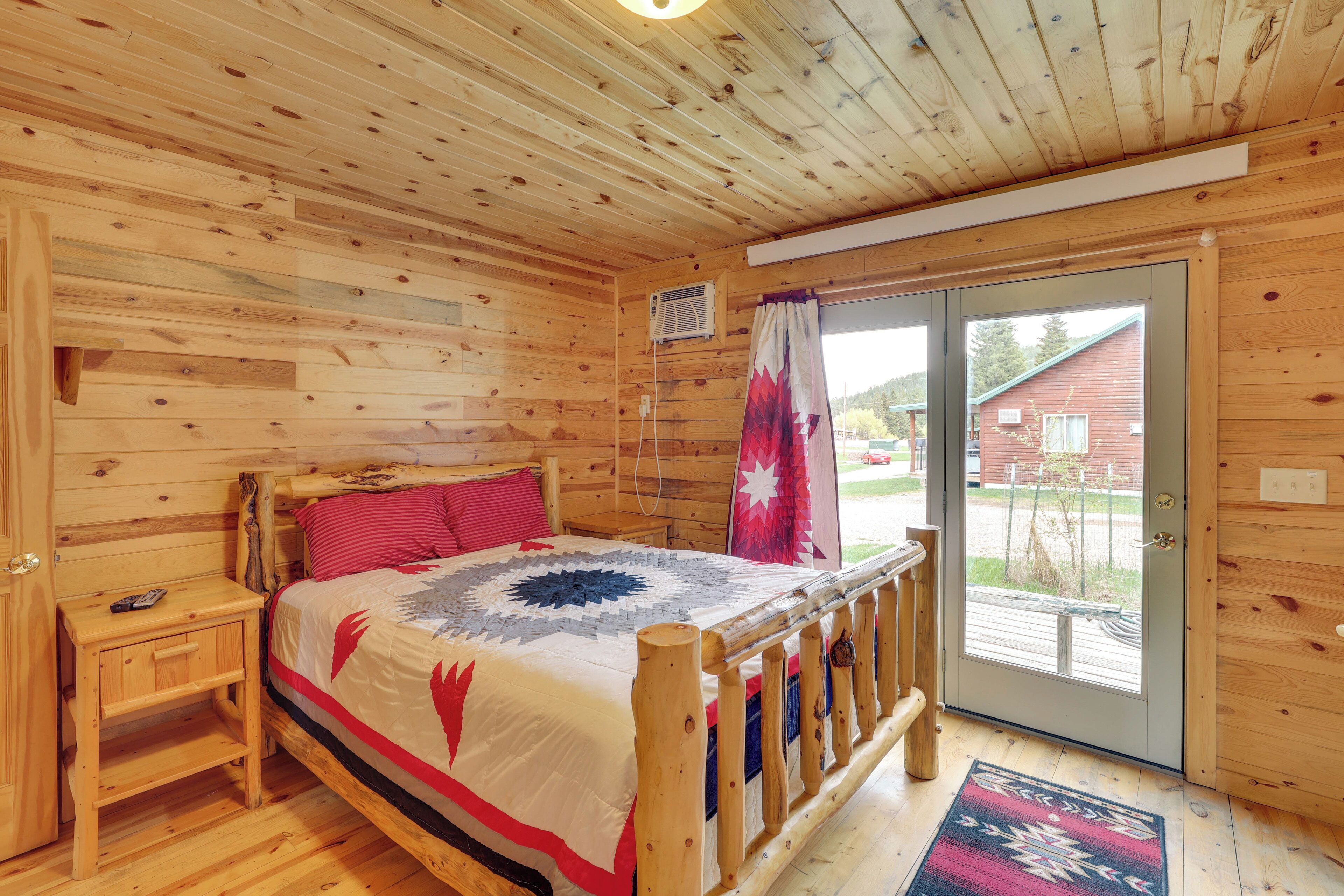 A wooden cabin bedroom with a bed, a nightstand, a window, and a door leading outside.