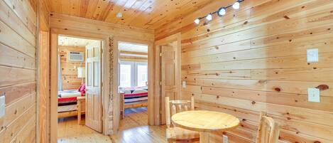 Interior de una cabaña de madera con comedor, una cama y un baño visible a través de una puerta abierta.