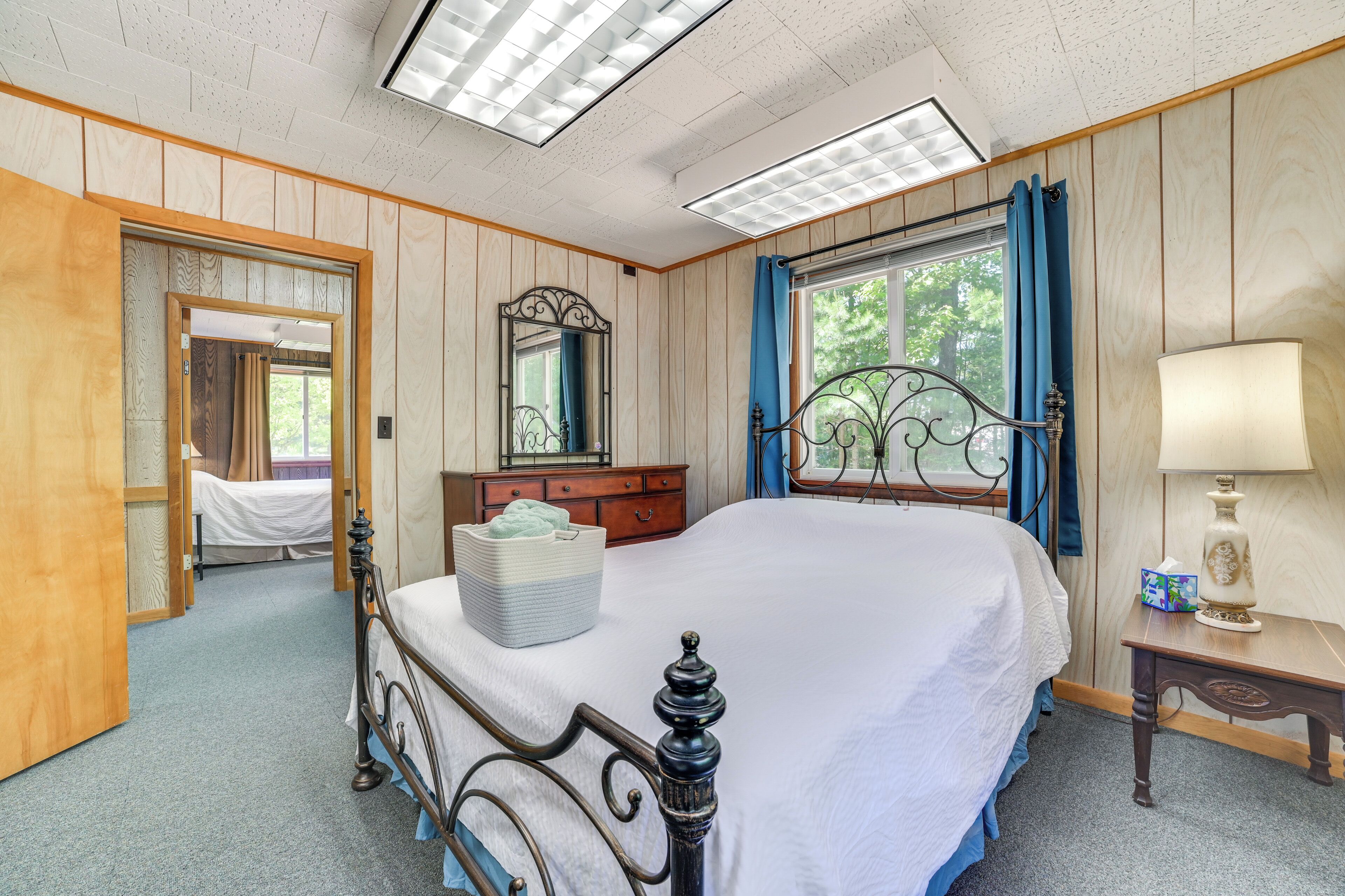 A bedroom with a large bed, a wooden dresser, a mirror, a window with curtains, and a bedside table with a lamp.
