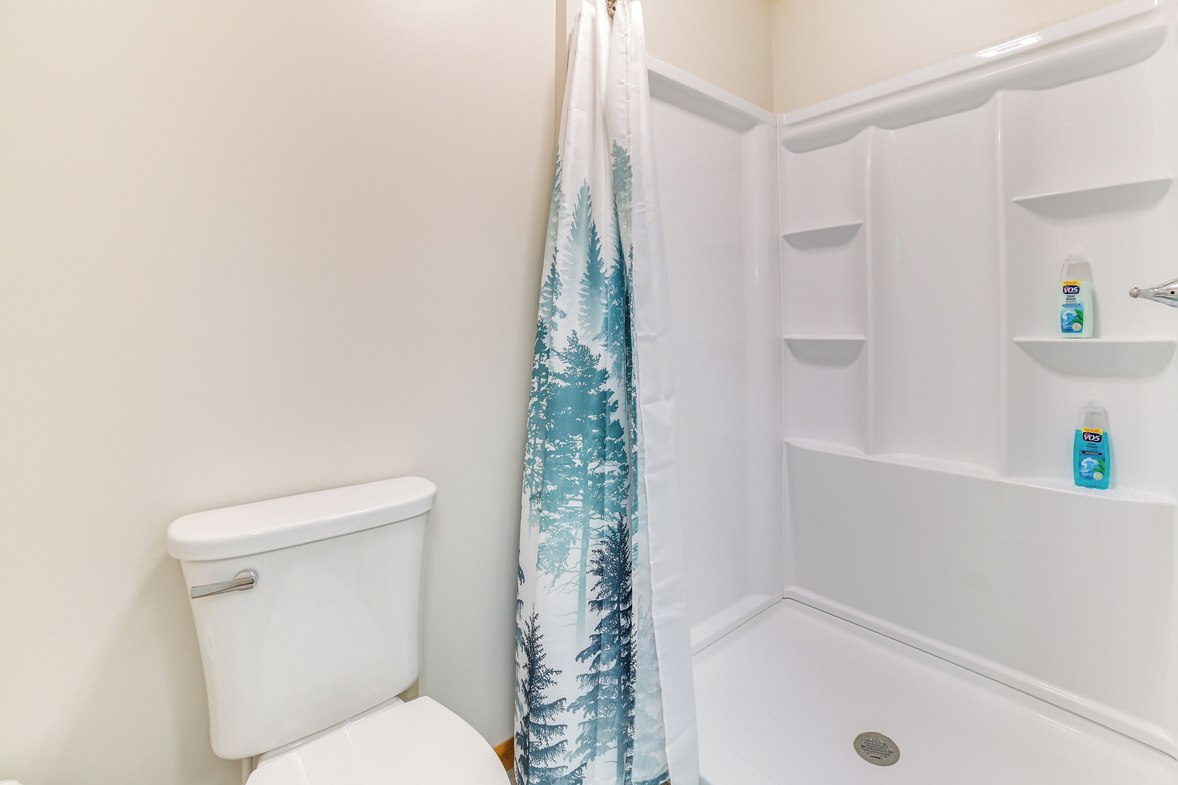 A bathroom with a white toilet, a shower with a blue forest-themed shower curtain, and a shelf with a bottle of hand soap.