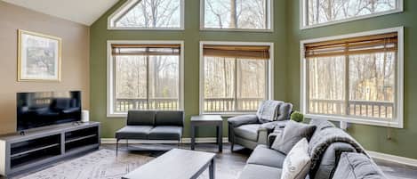 A living room with a large TV, two sofas, a coffee table, and large windows with blinds.