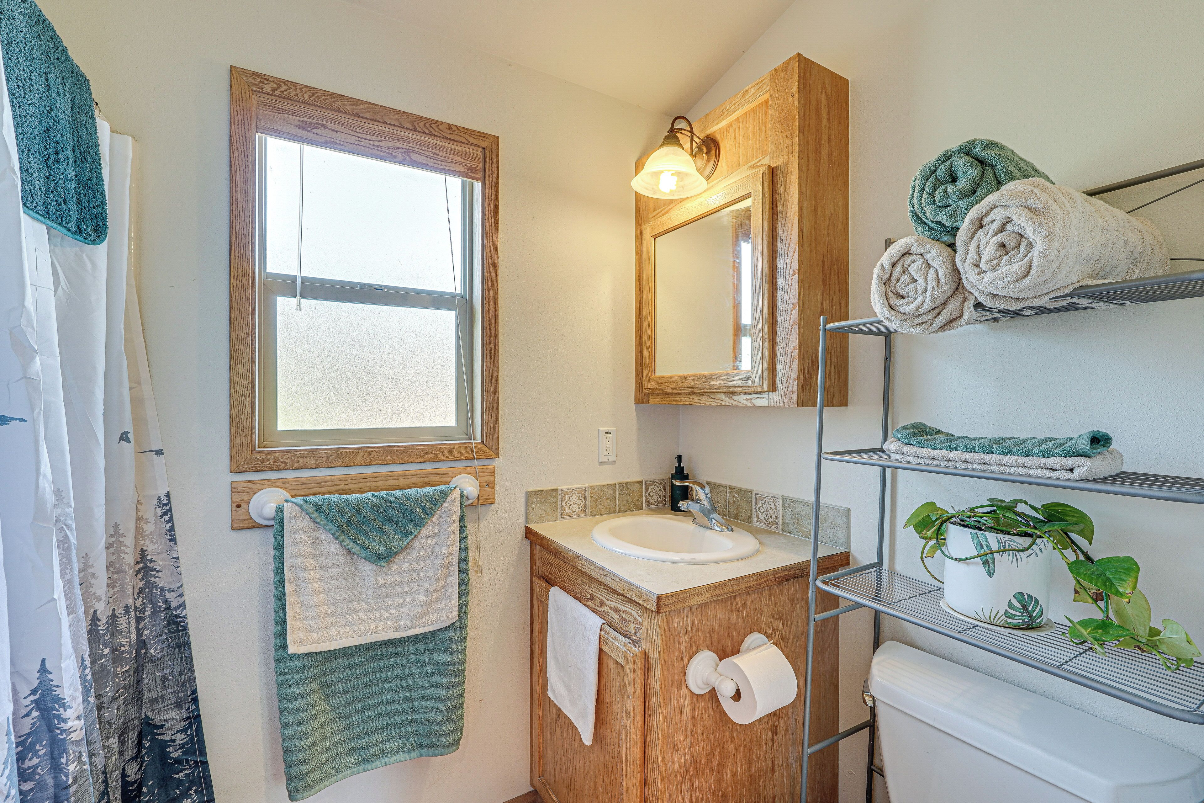 A bathroom with a wooden vanity, a mirror, a window, and shelves with towels and a potted plant.